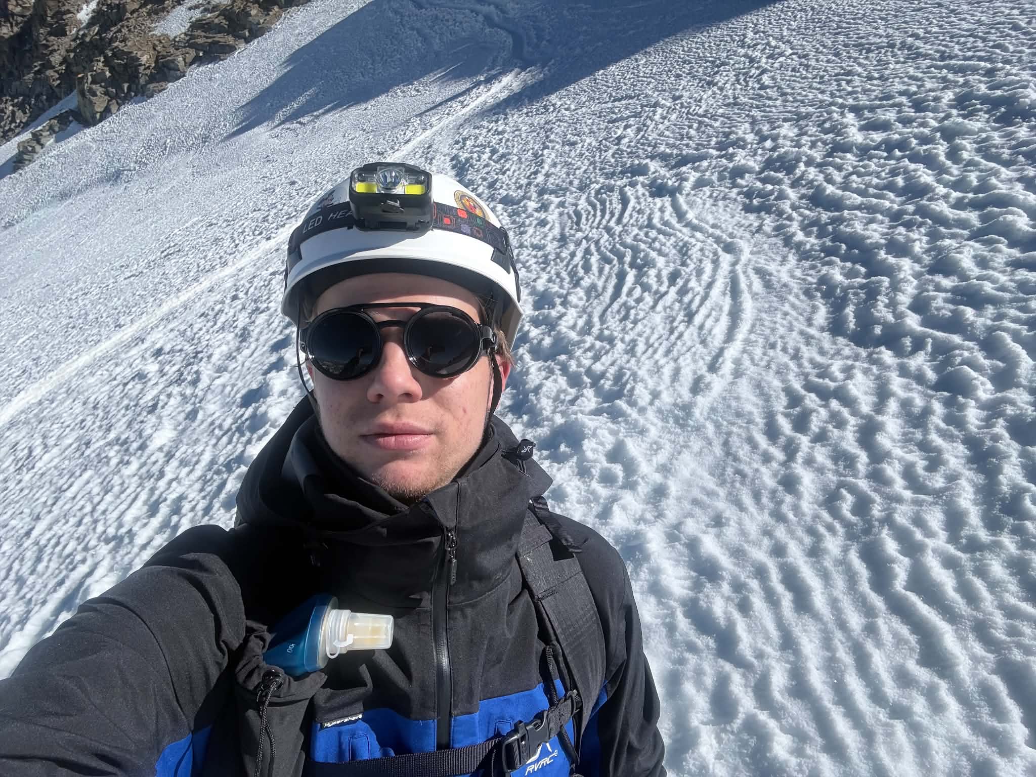 Selfie of Frank Andrew Annion in a white helmet with a headlamp and glacier glasses on a wind-rippled snow slope
