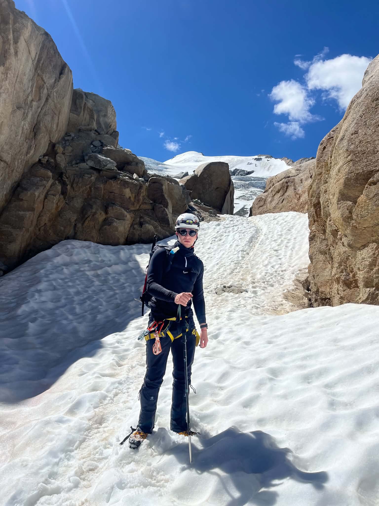 Frank Andrew Annion in a helmet and harness standing in a snow gully between granite walls below a glacier